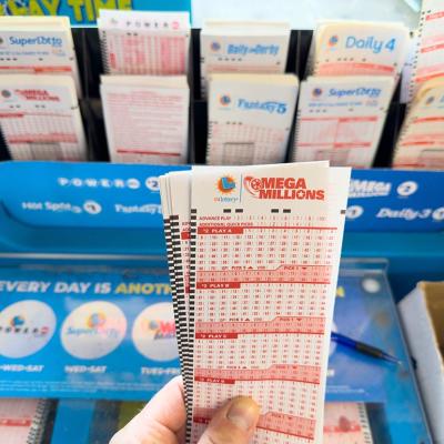 View of left hand holding Mega Millions tickets in front of a blue lottery kiosk.