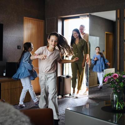 Happy family entering a hotel room with dark decor, parents in doorway with children running in foreground.