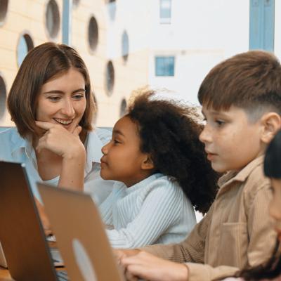 A female teacher looking at three young students while they all use laptops.