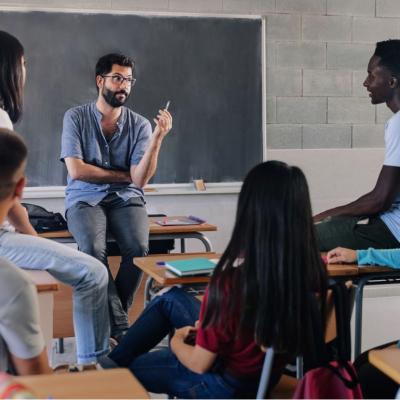 A male teacher sitting on top of the desk looking at the African American male student to his left in a classroom. The rest of the students are facing the teacher. 