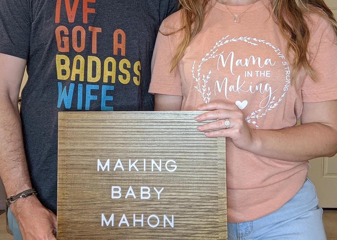 Self portrait of Kirsti and Justin Mahon from the neck down, holding a sign that says "Making Baby Mahon."