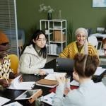 In an office, a diverse group of five young adults seated in a round table engaging in a discussion.