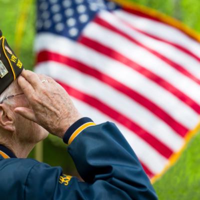 An older veteran saluting with an American flag waving in the background.