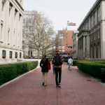 Students with backpacks walk with their backs to the camera on a college campus, surrounded by buildings.