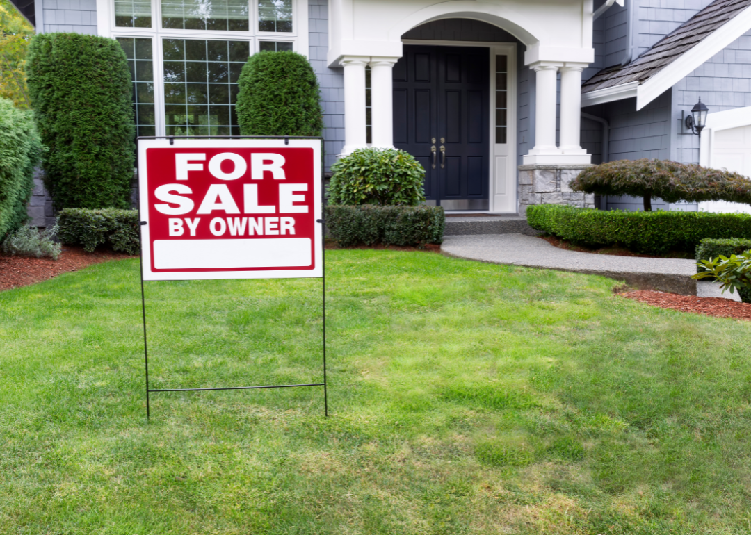 A for sale sign in the front yard of a suburban home.