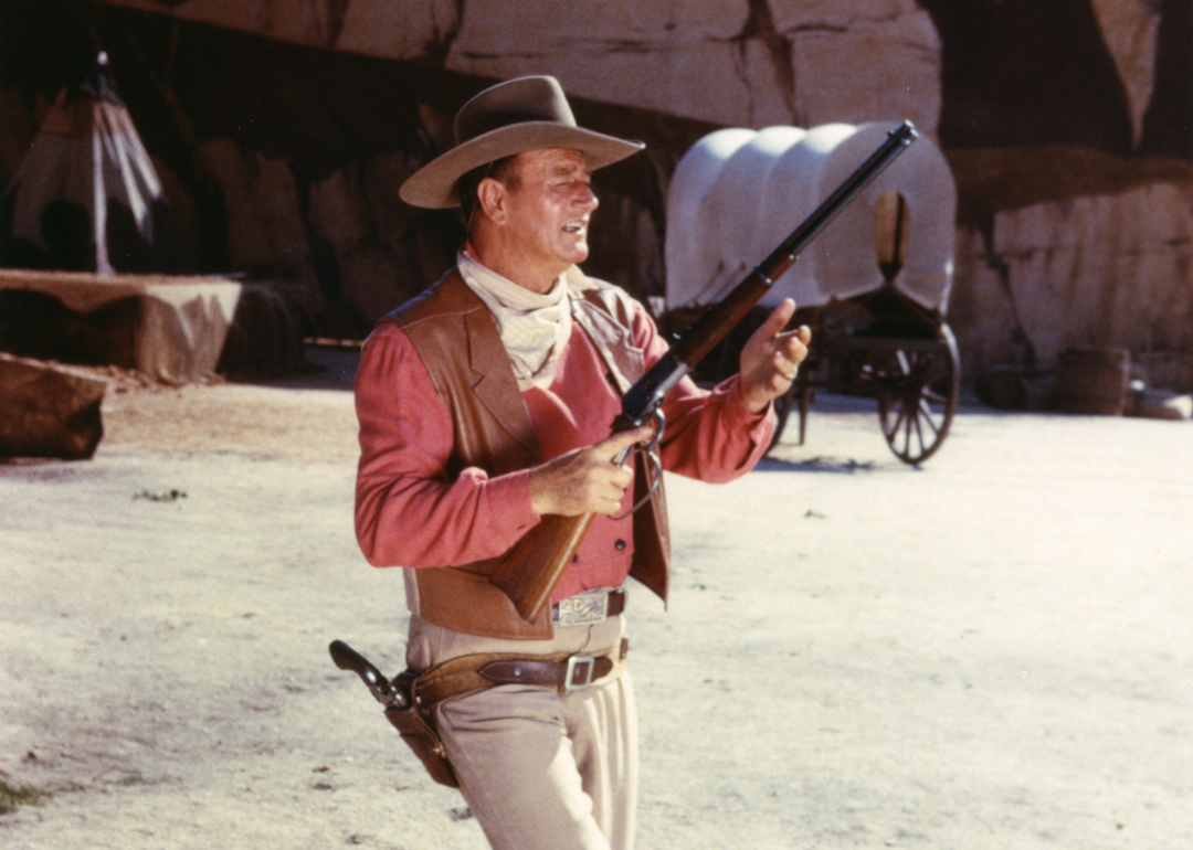 John Wayne (1907-1979), US actor, wearing a tan leather waistcoat over a pink shirt, taking aim with a rifle, in a publicity still issued for the film, 'Rio Bravo', USA, 1959. 