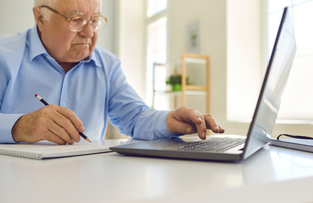 Senior man glancing at computer and taking notes in notebook.