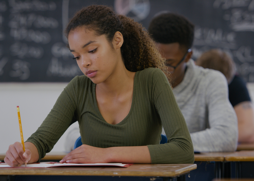 African American teenage girl sitting at the desk in a classroom holding a pencil and looking down at the paper in front of her.  