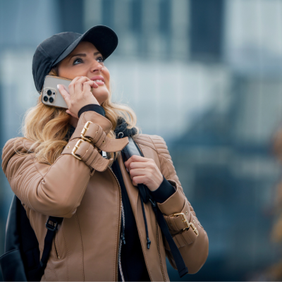 A woman in a black baseball hat and cream jacket holding a cellphone to her ear and looking up. 